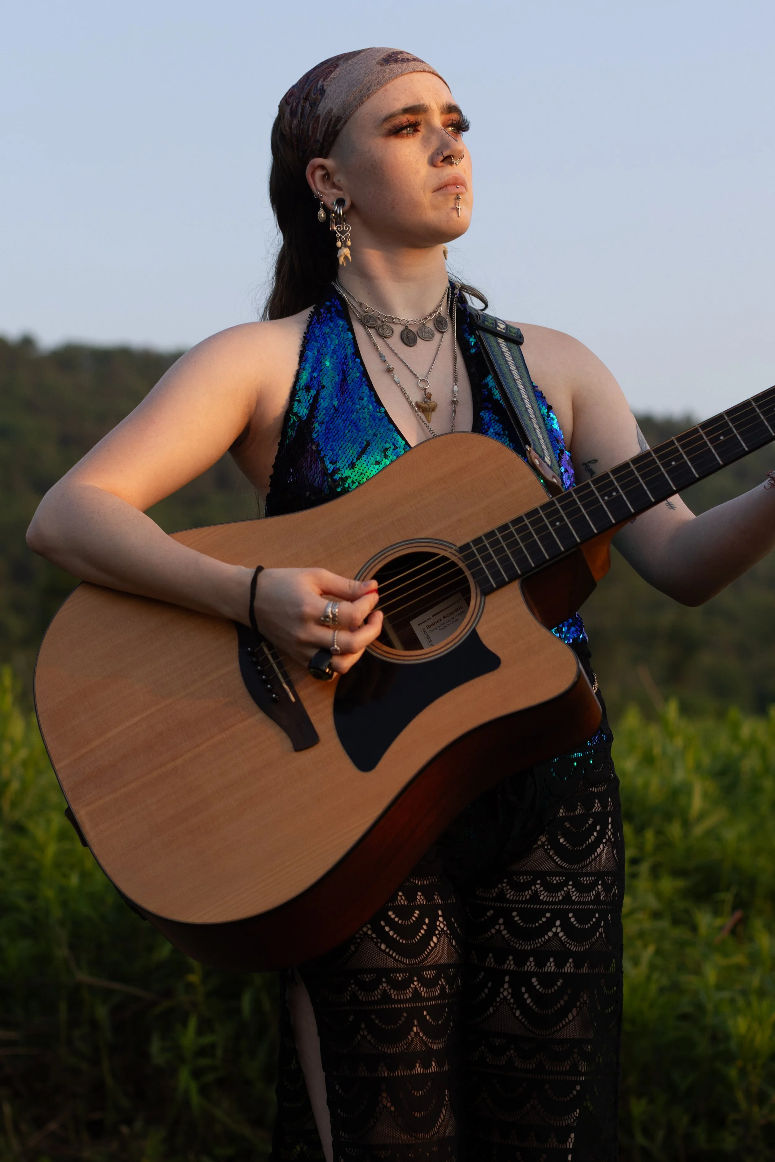 A young woman with tattoos and multiple jewelry pieces playing an acoustic guitar outdoors during sunset, wearing a sleeveless blue and black top and black lace pants.