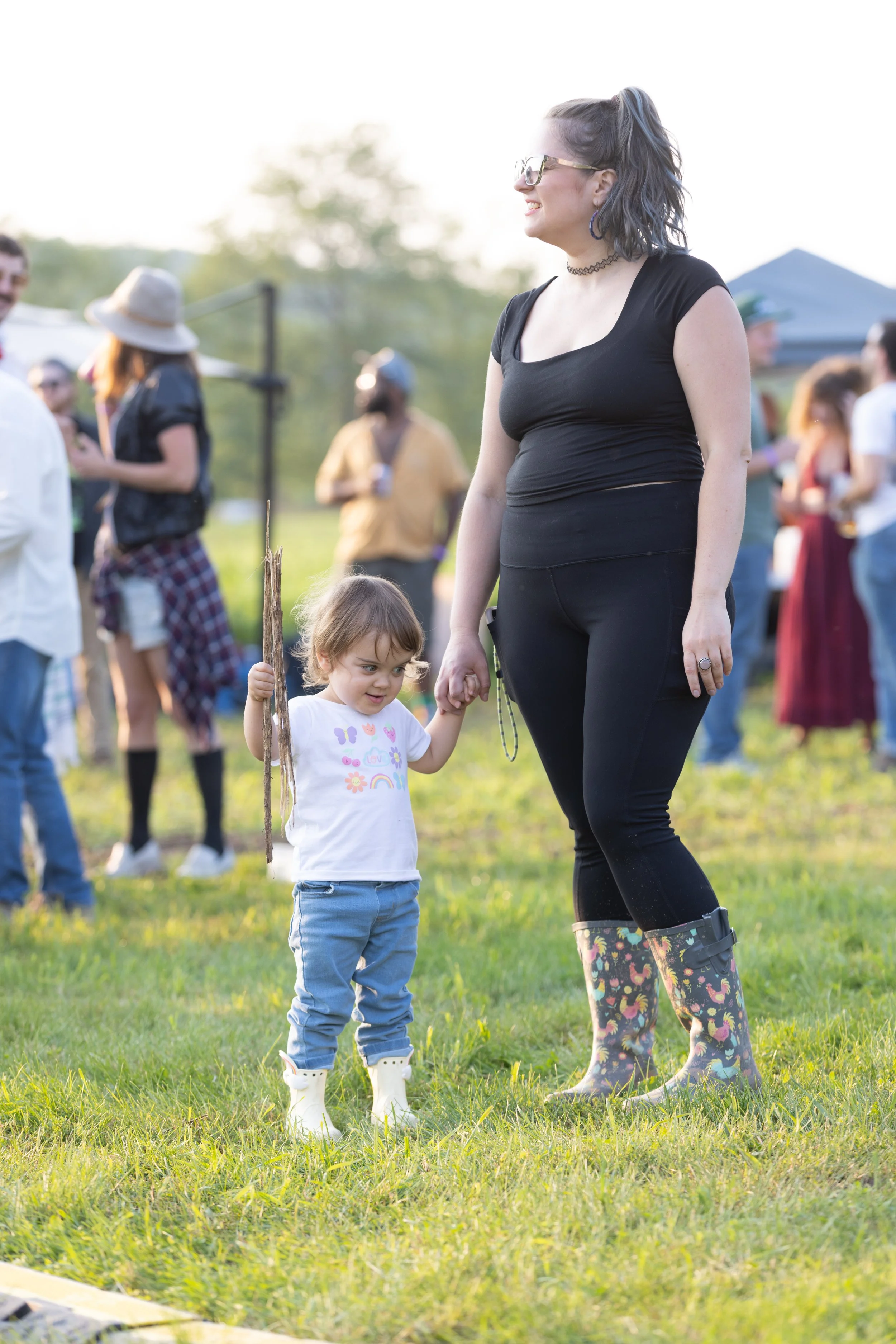 A woman with glasses, a black shirt, and patterned leggings holds hands with a young girl wearing a white shirt, blue jeans, and rain boots, outdoors in a grassy area with other people in the background.