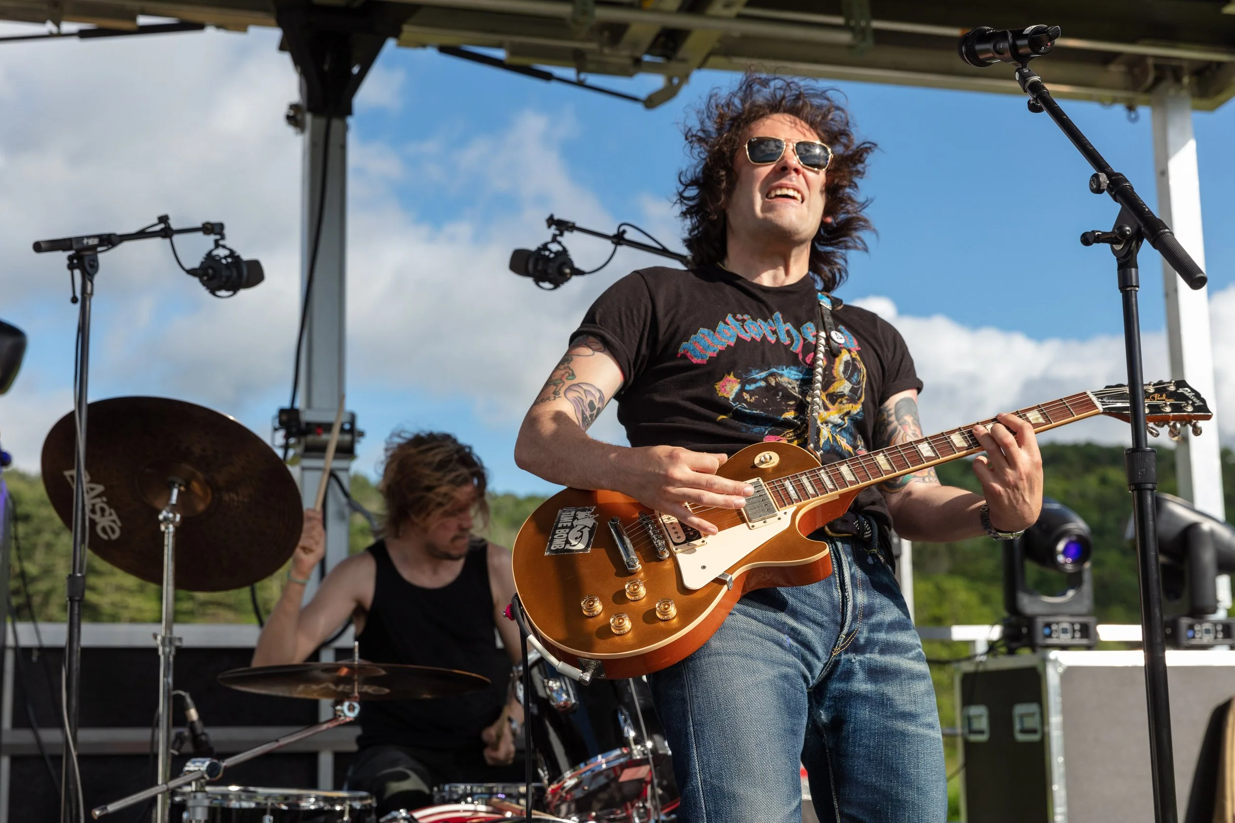 Musician with curly hair wearing sunglasses and a black T-shirt playing an electric guitar on an outdoor stage with a blue sky background.