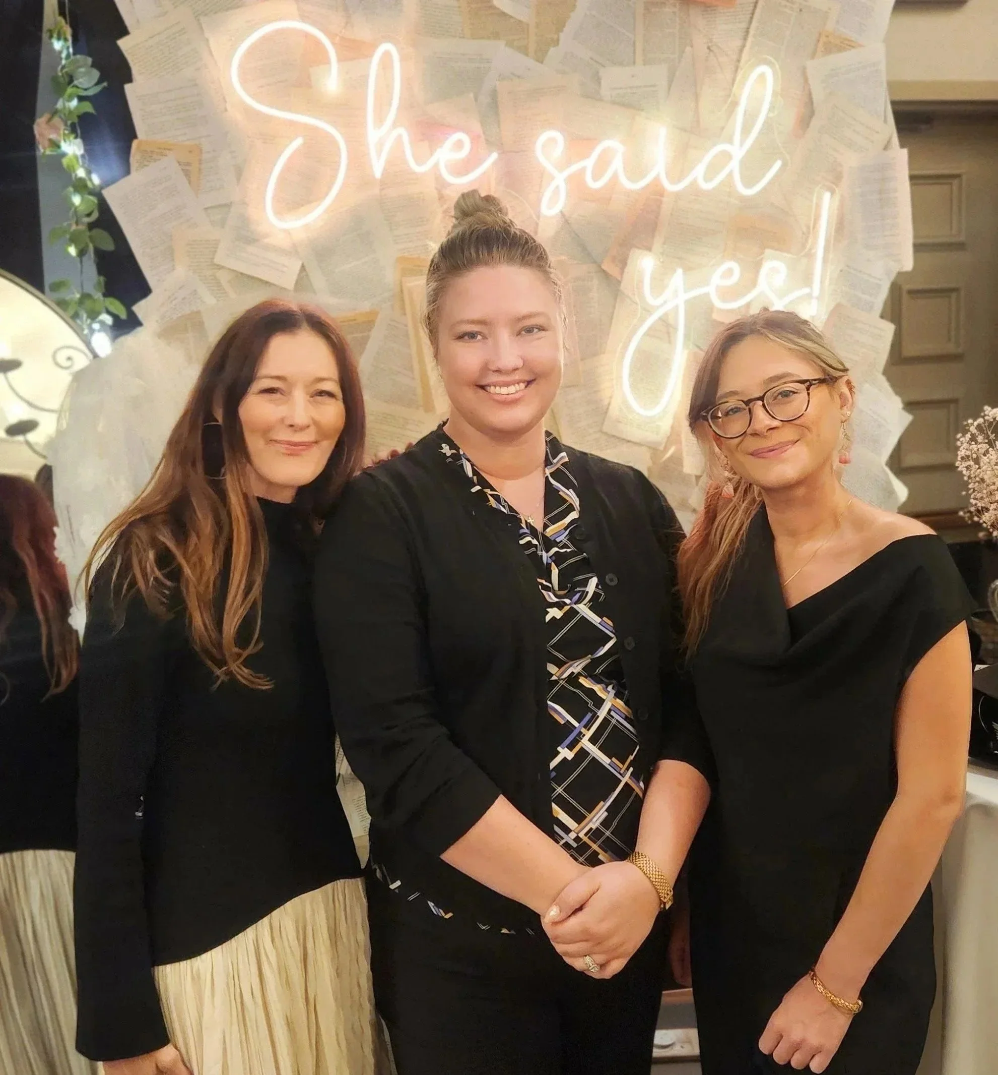 Three women standing together in front of a wall decorated with papers and a neon sign that reads 'She said yes!'. They are smiling and appear happy. The woman in the center is wearing a black outfit with a patterned collar, the woman on the left is in a black top and a beige pleated skirt, and the woman on the right is wearing glasses and a black top.