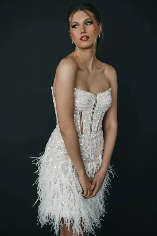 A woman in a strapless white wedding gown with lace details, standing in a rustic room with wooden floors and brick walls.