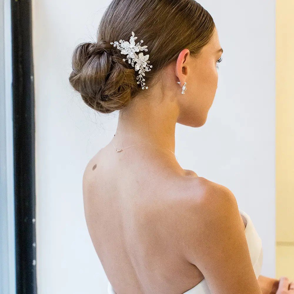 Side view of a woman with brunette hair styled in a low bun, adorned with a decorative hairpiece, wearing pearl earrings and a necklace, dressed in a strapless wedding gown.