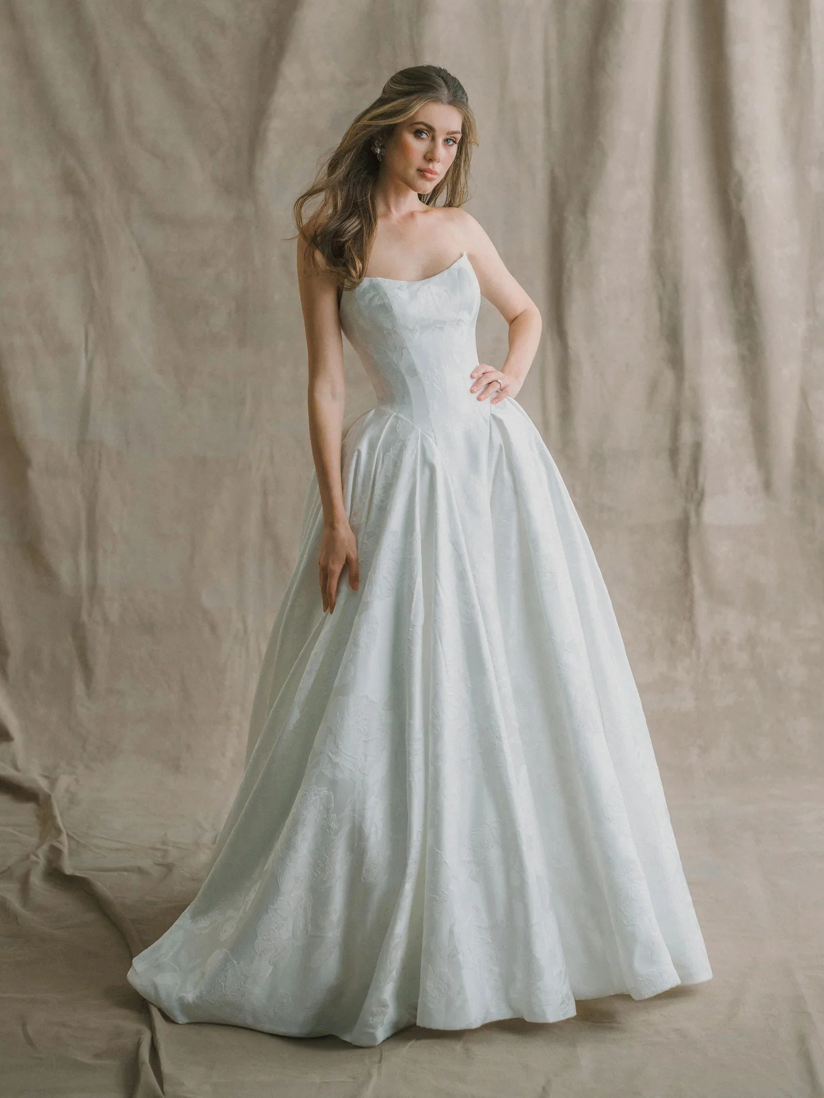 A woman in a strapless white wedding gown with lace details, standing in a rustic room with wooden floors and brick walls.