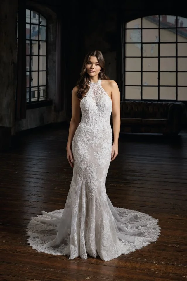 A woman in a white strapless wedding gown with lace details, standing against a dark background.