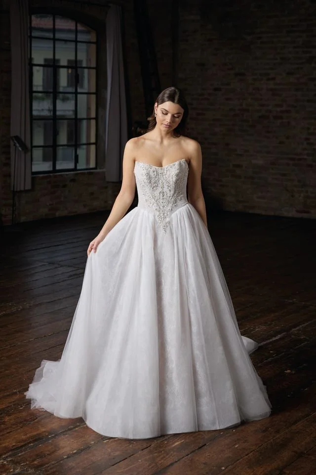 A woman in a white wedding dress with floral lace details is walking up an outdoor stone staircase in a European-style neighborhood. She is smiling and looking down, holding her dress slightly lifted to reveal beige high-heeled shoes. She has her hair pulled back and is wearing large earrings.