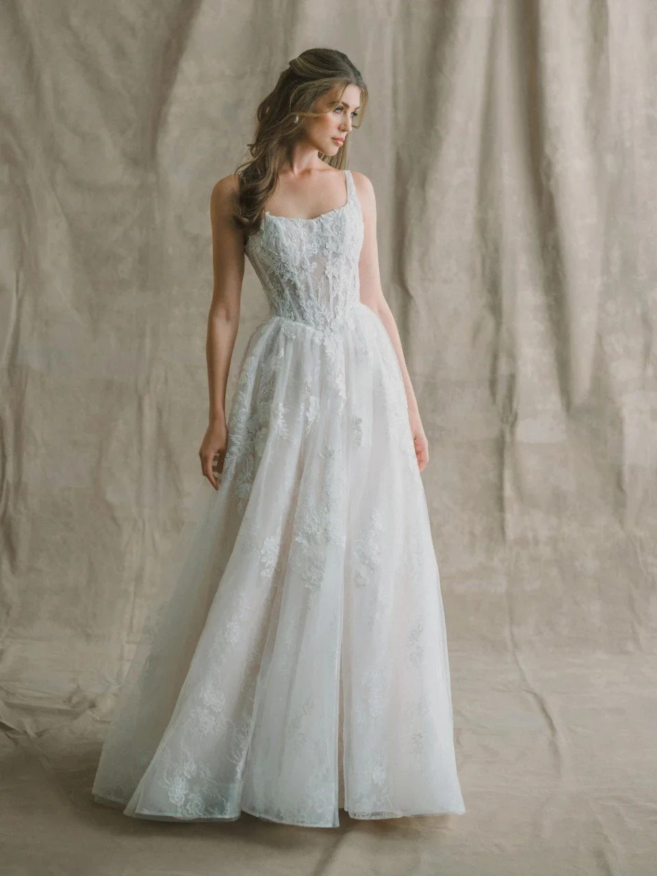 A woman in a white strapless wedding gown with lace details, standing against a dark background.