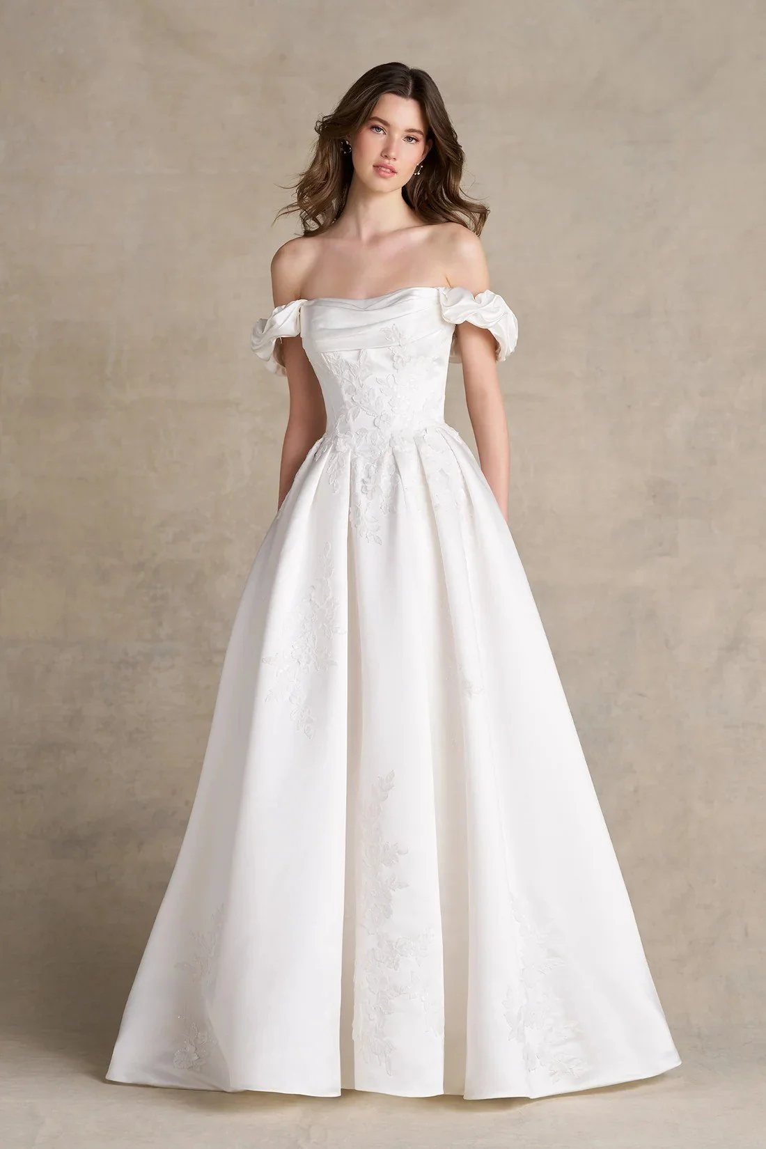 A woman wearing a white strapless wedding dress with floral embroidery, standing against a plain gray background.