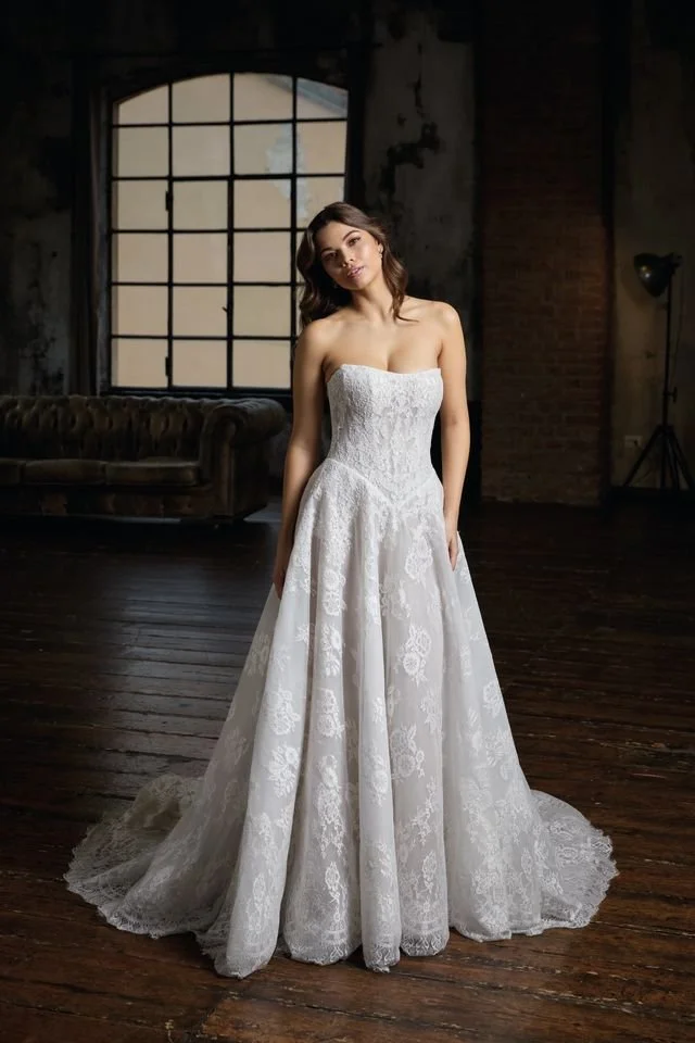 A woman wearing a white strapless wedding dress with floral embroidery, standing against a plain gray background.