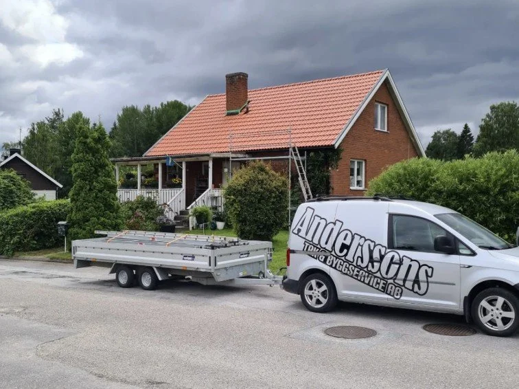 En vit servicebil med texten 'Andersson' parkerad framför ett rött hus med täckt veranda och trädgård, med ett tomt släp bakom.
