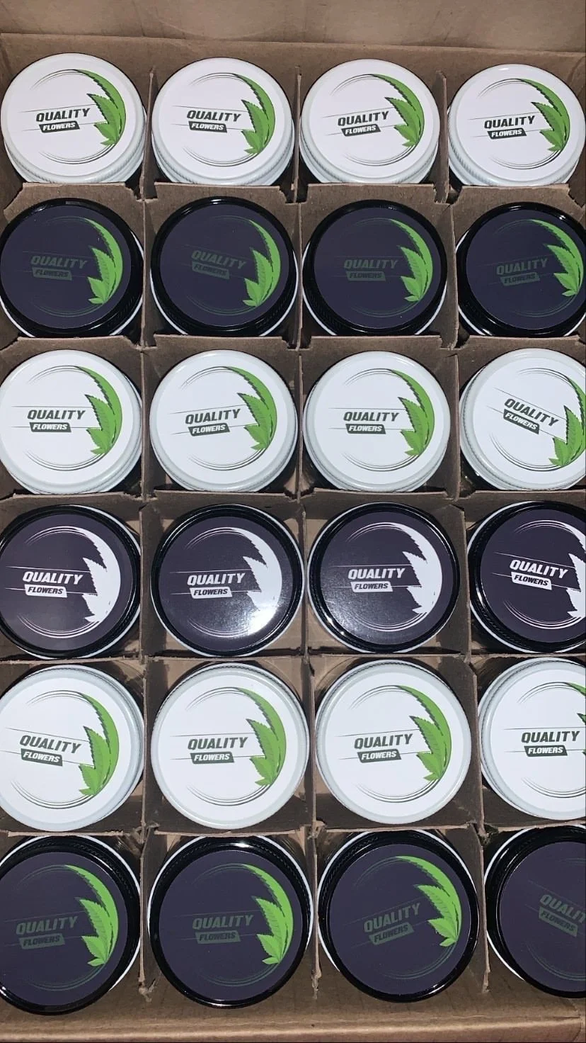 A display box of small round containers labeled "Quality Flowers," with alternating black and white lids featuring a green leaf logo.