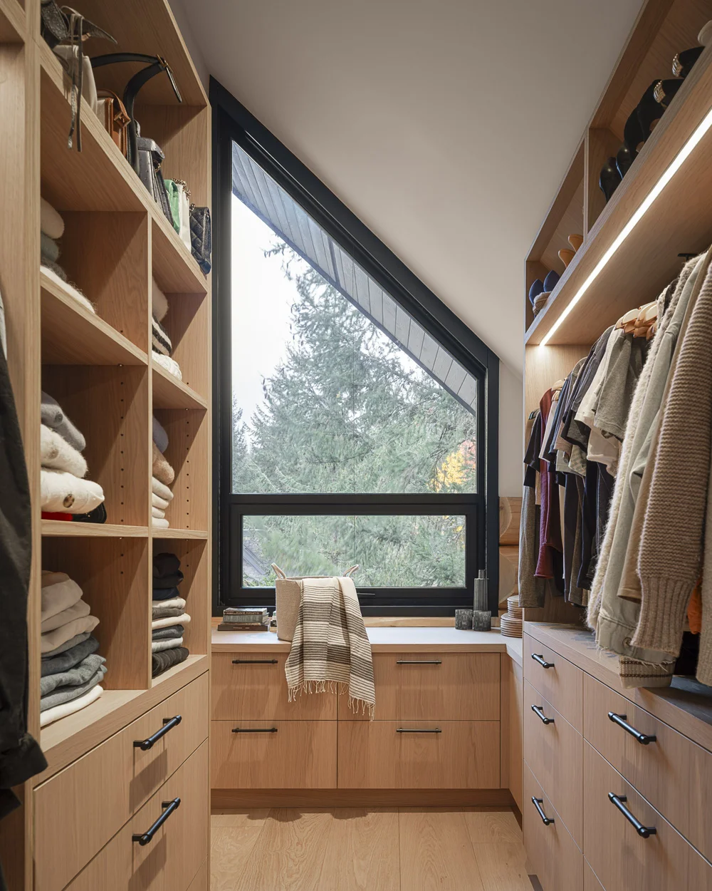 Walk in closet under a sloped ceiling, folded knits on open shelves beside a window