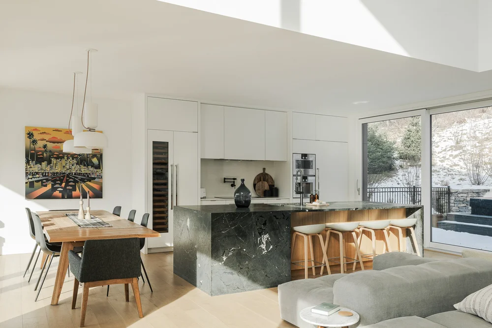 Open plan room with a dining table, a dark stone island and glazing onto a snowy slope