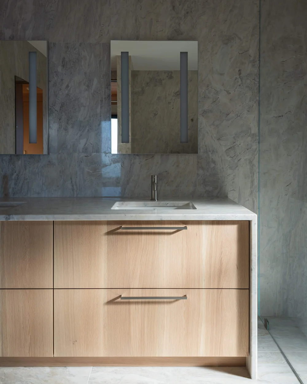 Pale wood vanity under a stone counter, two mirrors set against the stone wall