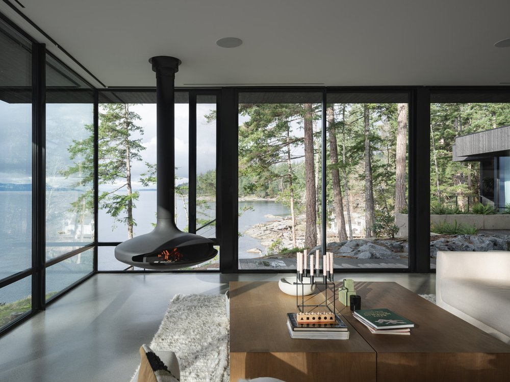 Living room corner with a lit suspended fireplace, glazing onto the water and rocky shore