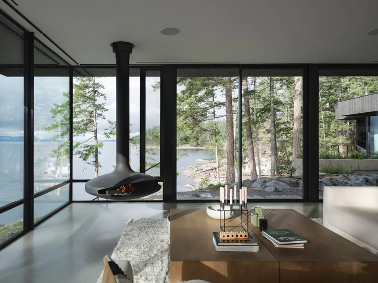 Living room corner with a lit suspended fireplace, glazing onto the water and rocky shore