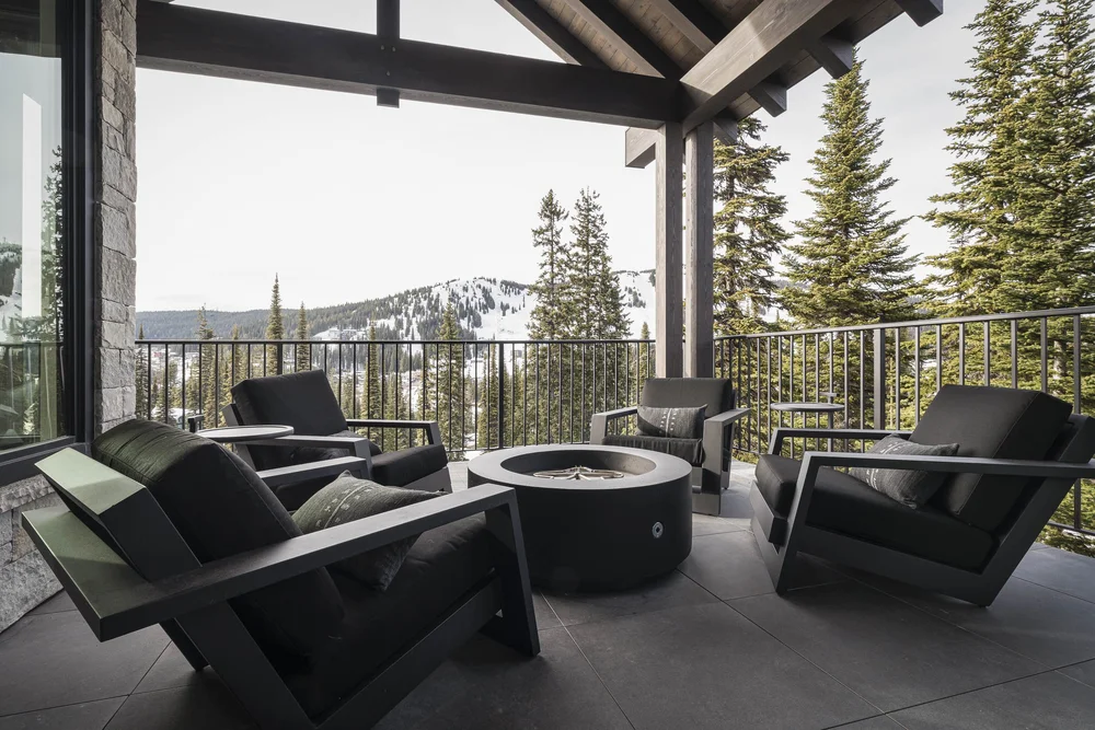 Covered deck with dark chairs around a round fire table, ski runs beyond