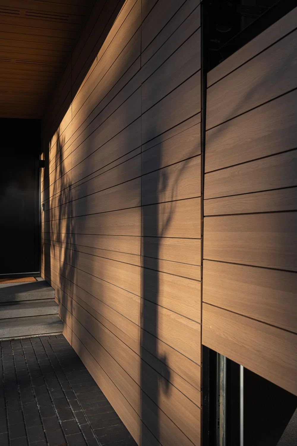 Looking along the wood clad entry wall, shadows raking the boards above a dark walkway