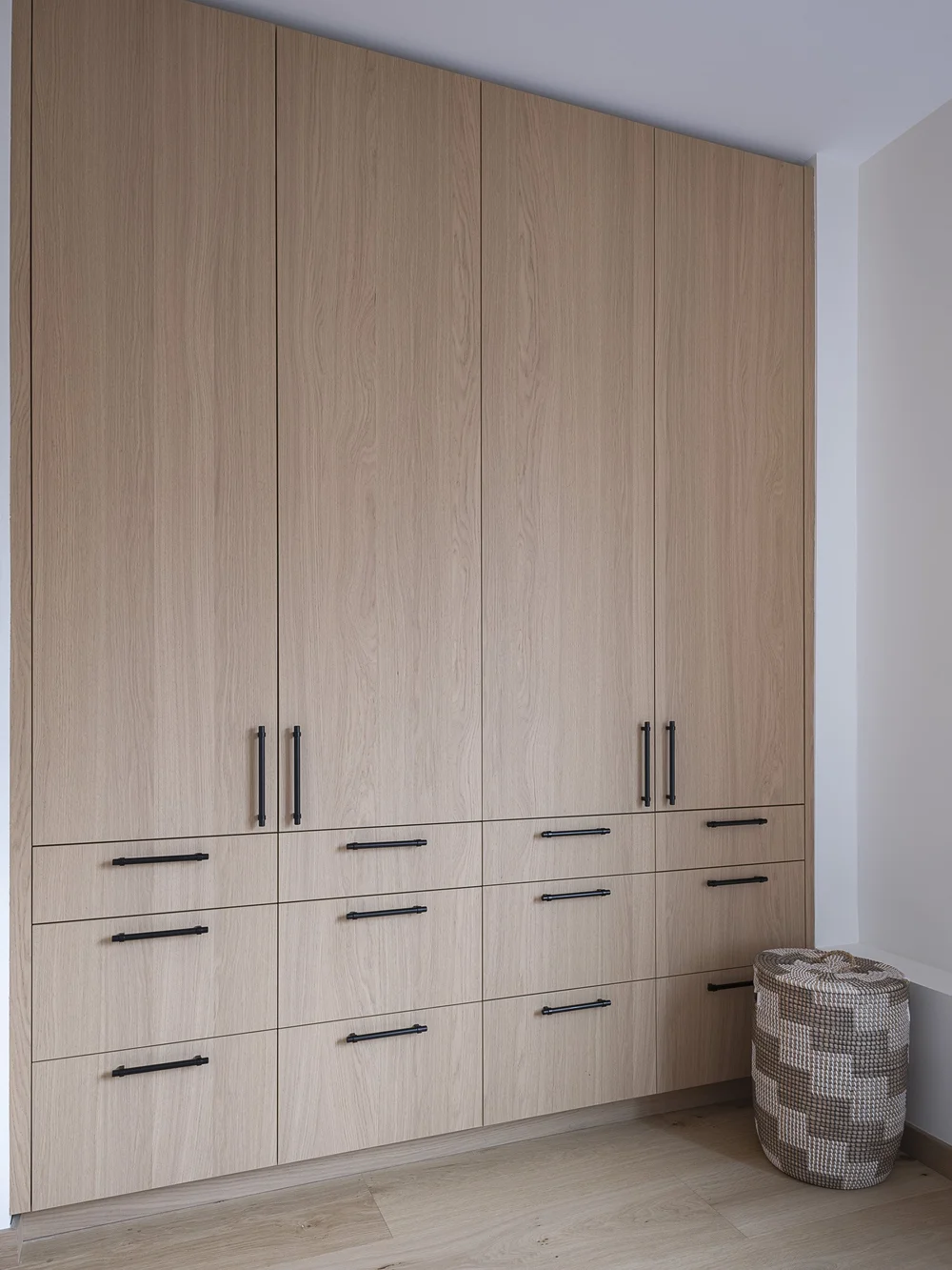 Full height wardrobes in pale wood with black bar pulls, a woven basket alongside