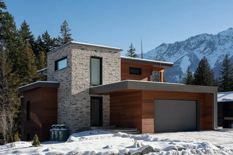 The house in snow below a mountain ridge, a stone volume beside horizontal wood cladding