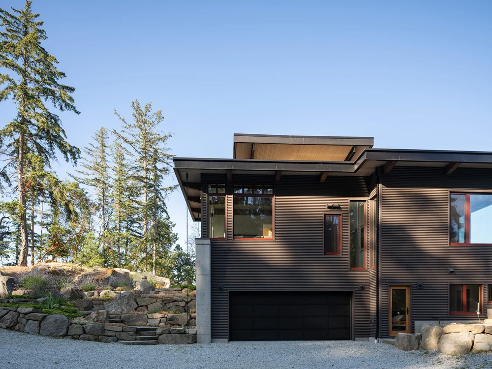 Garage wing in dark ribbed cladding above a boulder retaining wall and gravel drive