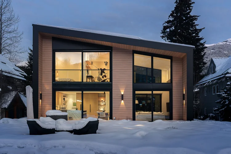 The house at dusk, interiors lit behind tall glazing with snow across the terrace