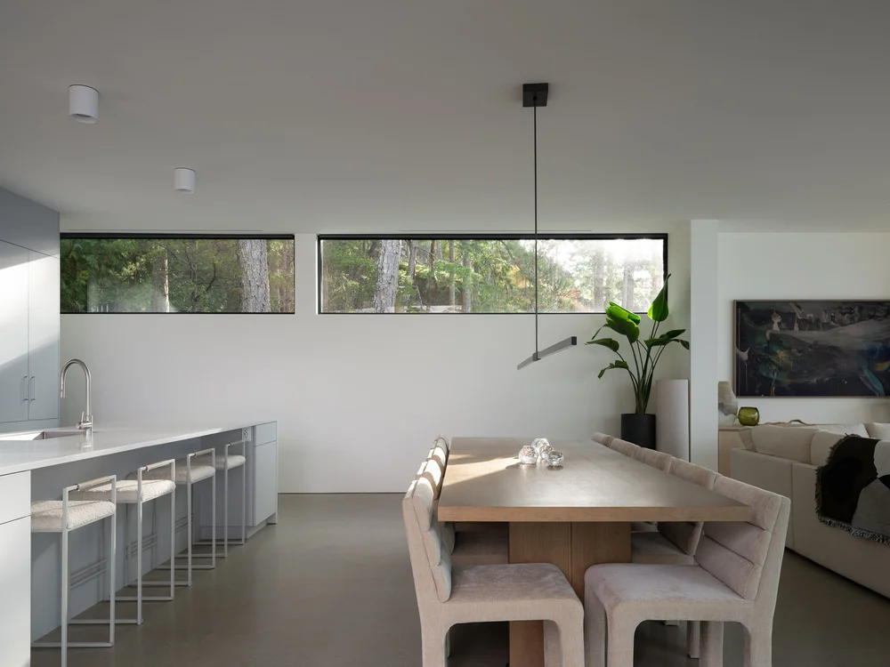 Kitchen island and stools beside a long dining table, ribbon windows onto the trees