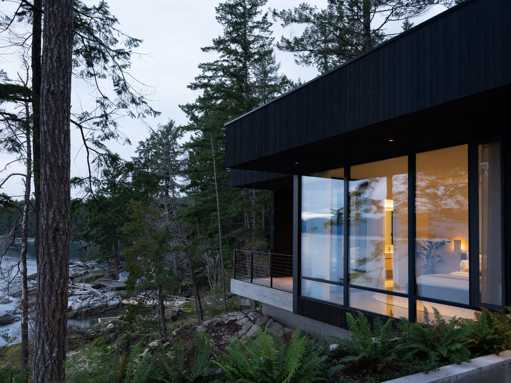 Dusk, a lit bedroom behind glass above a concrete base with ferns across the rock
