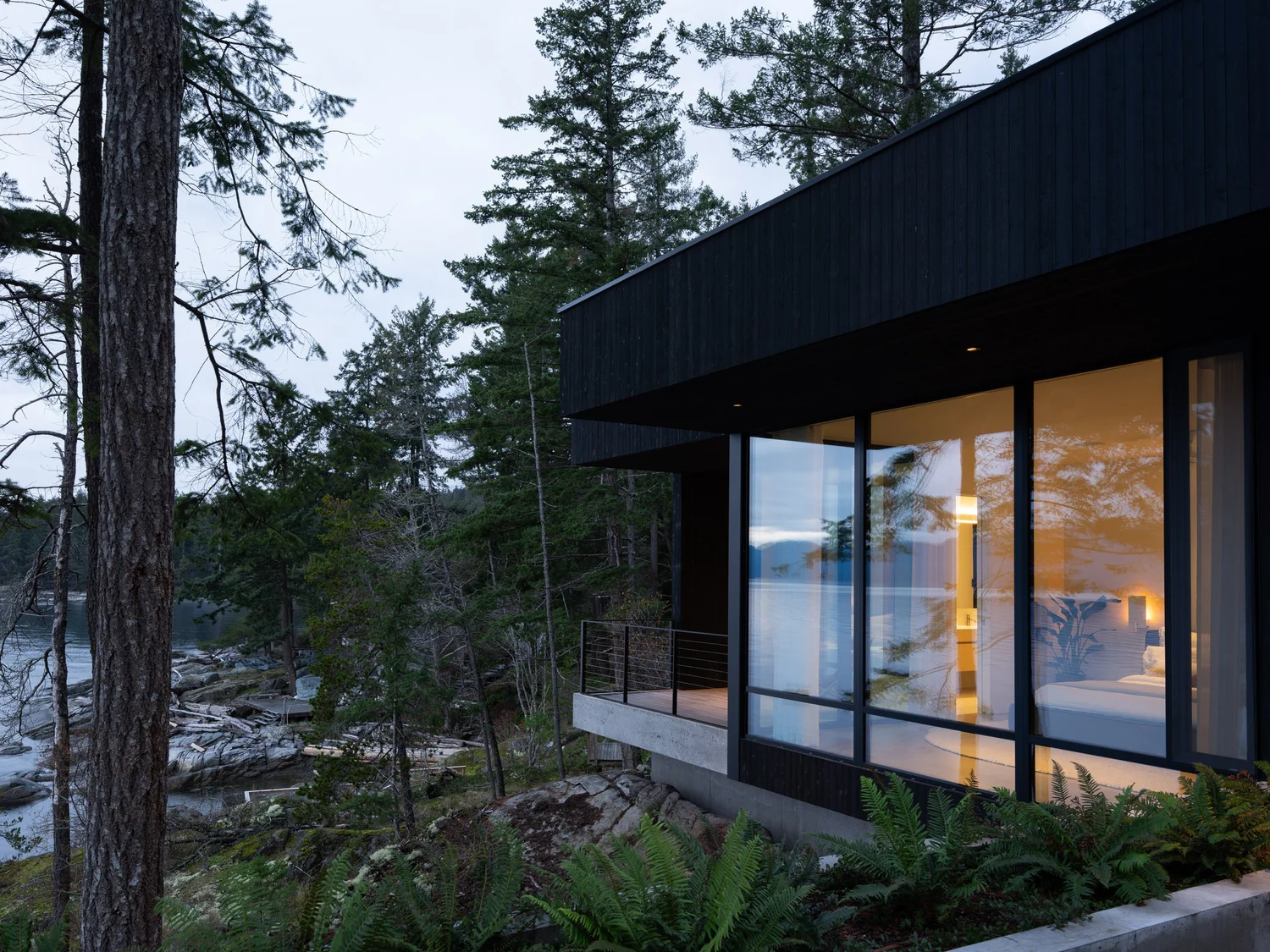 Dusk, a lit bedroom behind glass above a concrete base with ferns across the rock