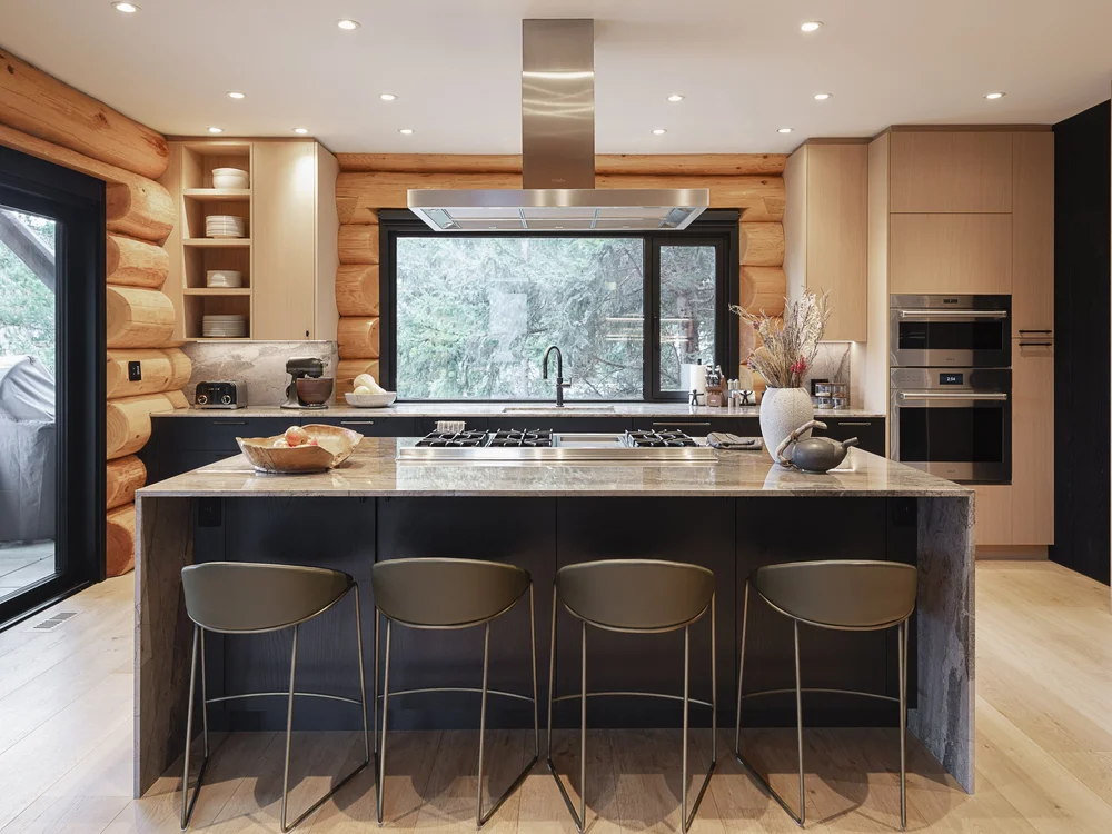Island with a veined stone top and four stools, wall ovens and pale cabinetry behind
