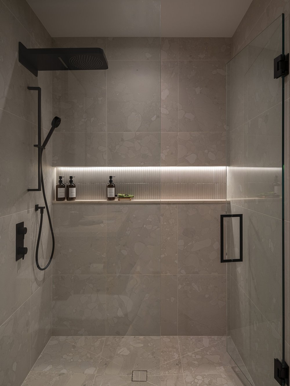 Walk in shower in large tile, black rain head above a lit niche shelf