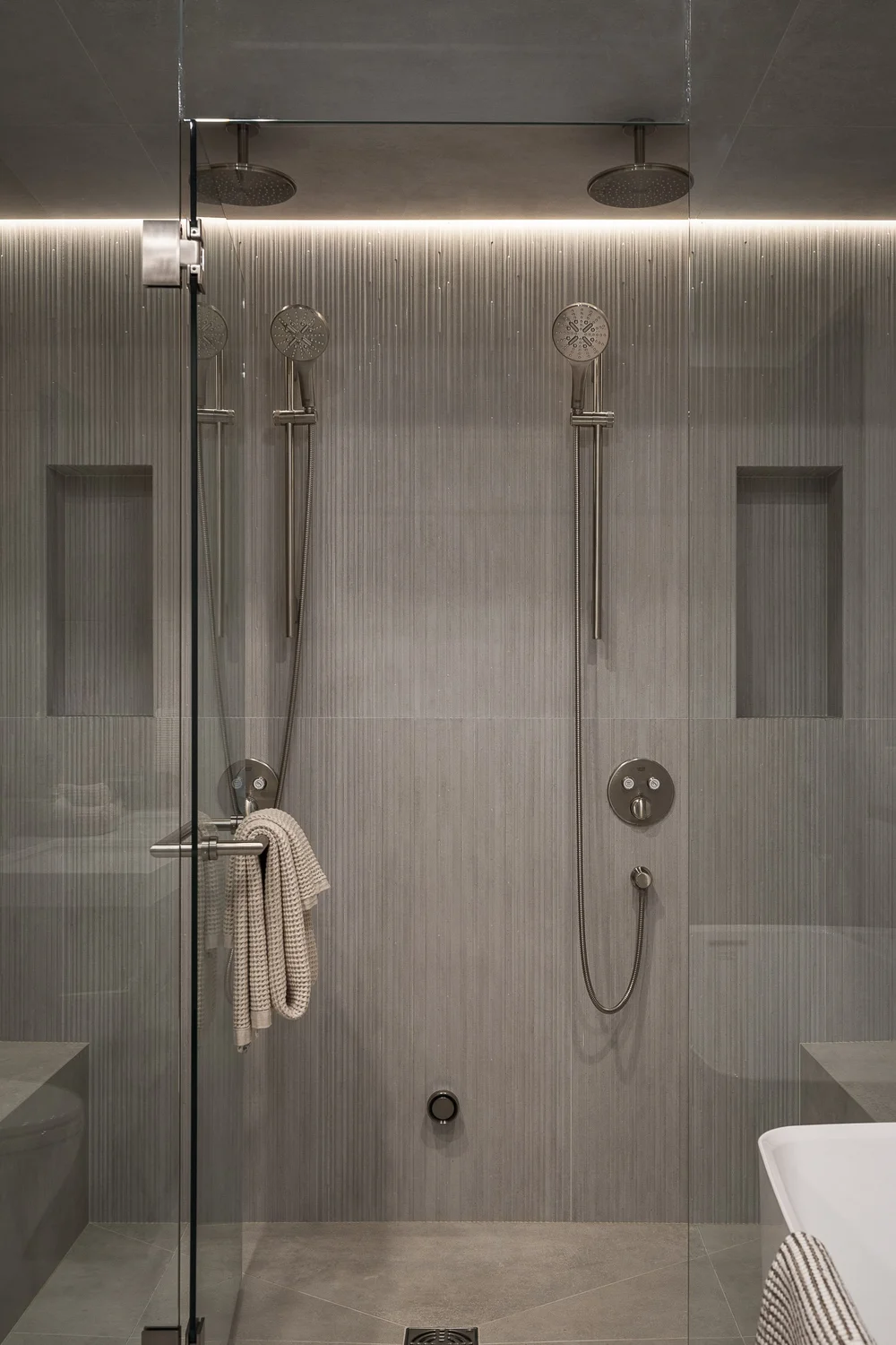 Two rainheads and handshowers in a tiled shower behind clear glass, a towel hanging