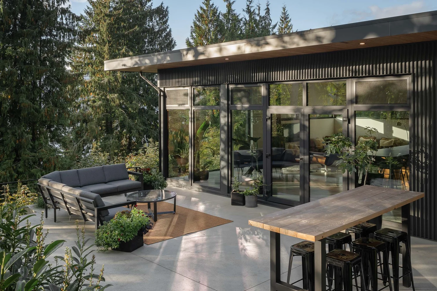 Glazed elevation opening to a concrete terrace, outdoor seating and a wood topped bar under tall firs