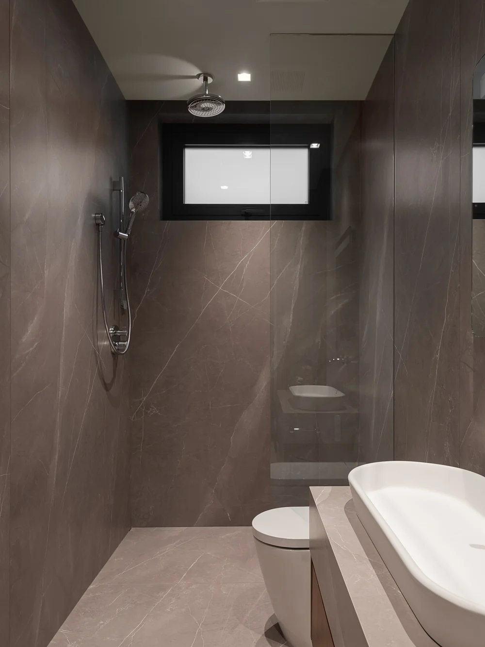 Shower in dark stone tile with a slot window, a toilet and tub at right