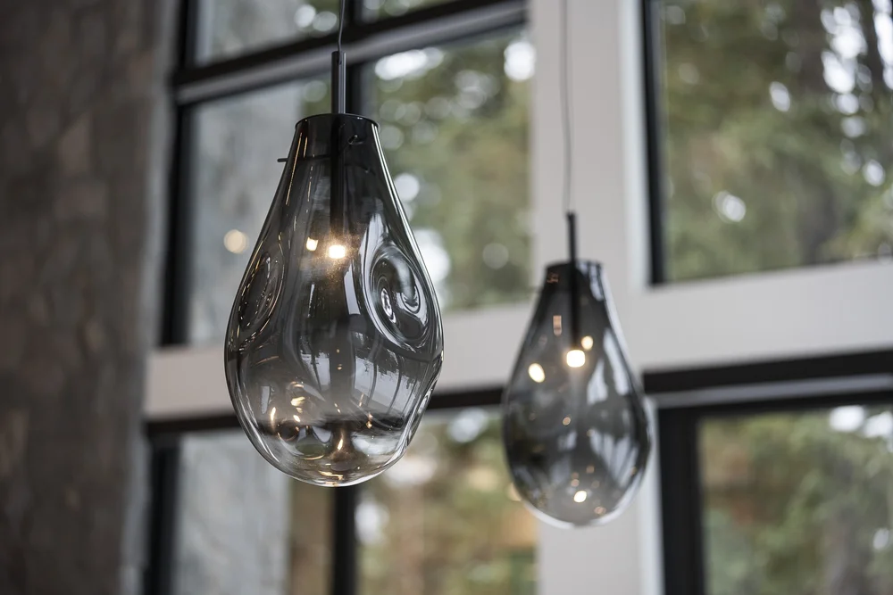 Two smoked glass pendants hanging in front of tall black framed glazing and trees