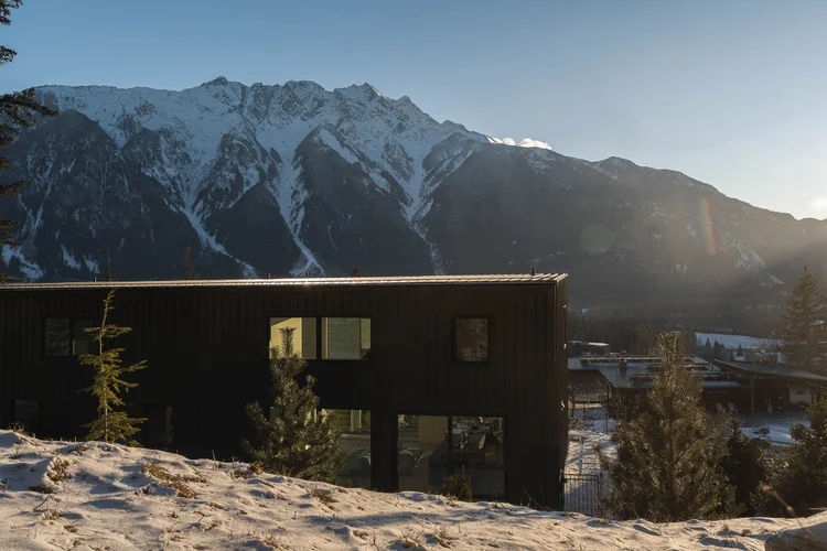 The house low on a snowy slope, a long dark elevation with snow covered peaks beyond