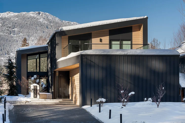Dark metal and cedar entry elevation in snow with a mountain slope behind