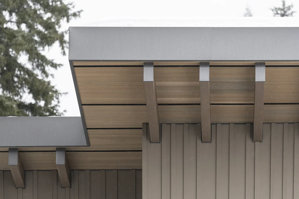 Two roof planes meeting over vertical siding, beam tails exposed, snow along the lower edge