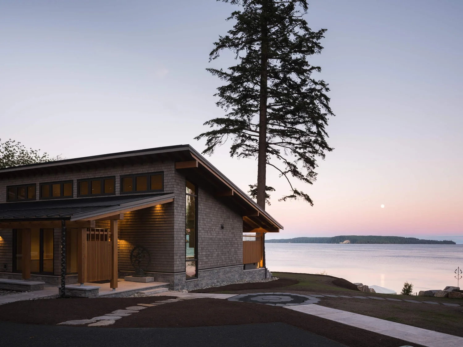 The house at dusk above the shore, the entry lit and the moon over the water
