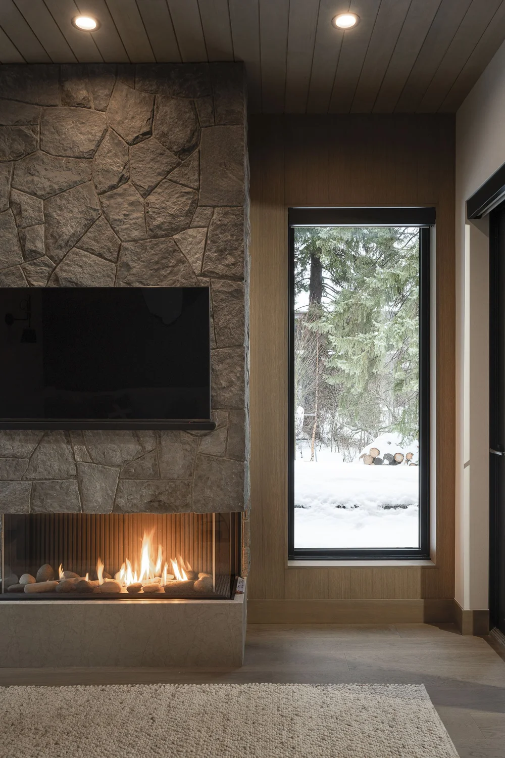 Stone clad fireplace with a long fire behind glass and a screen above, tall window at right