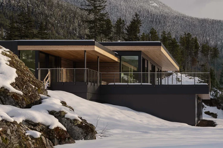 The house along a rock outcrop above the snow, a long dark volume under a pale wood soffit