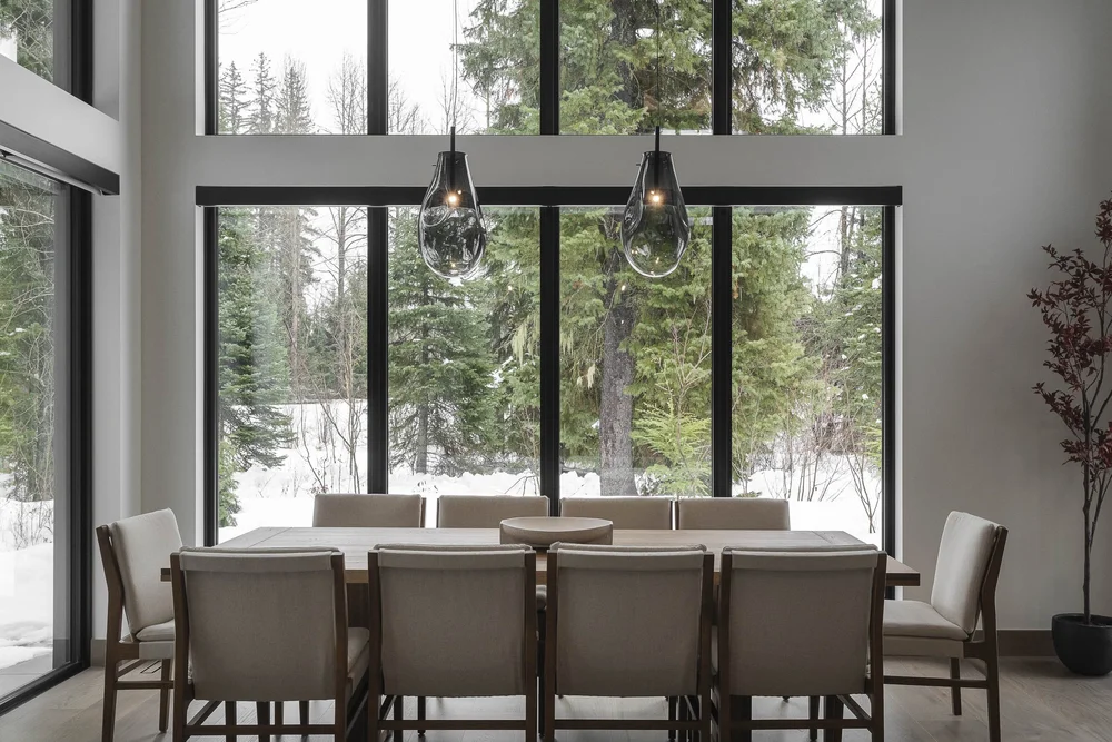Dining table and chairs under two glass pendants, full height glazing to snow and conifers