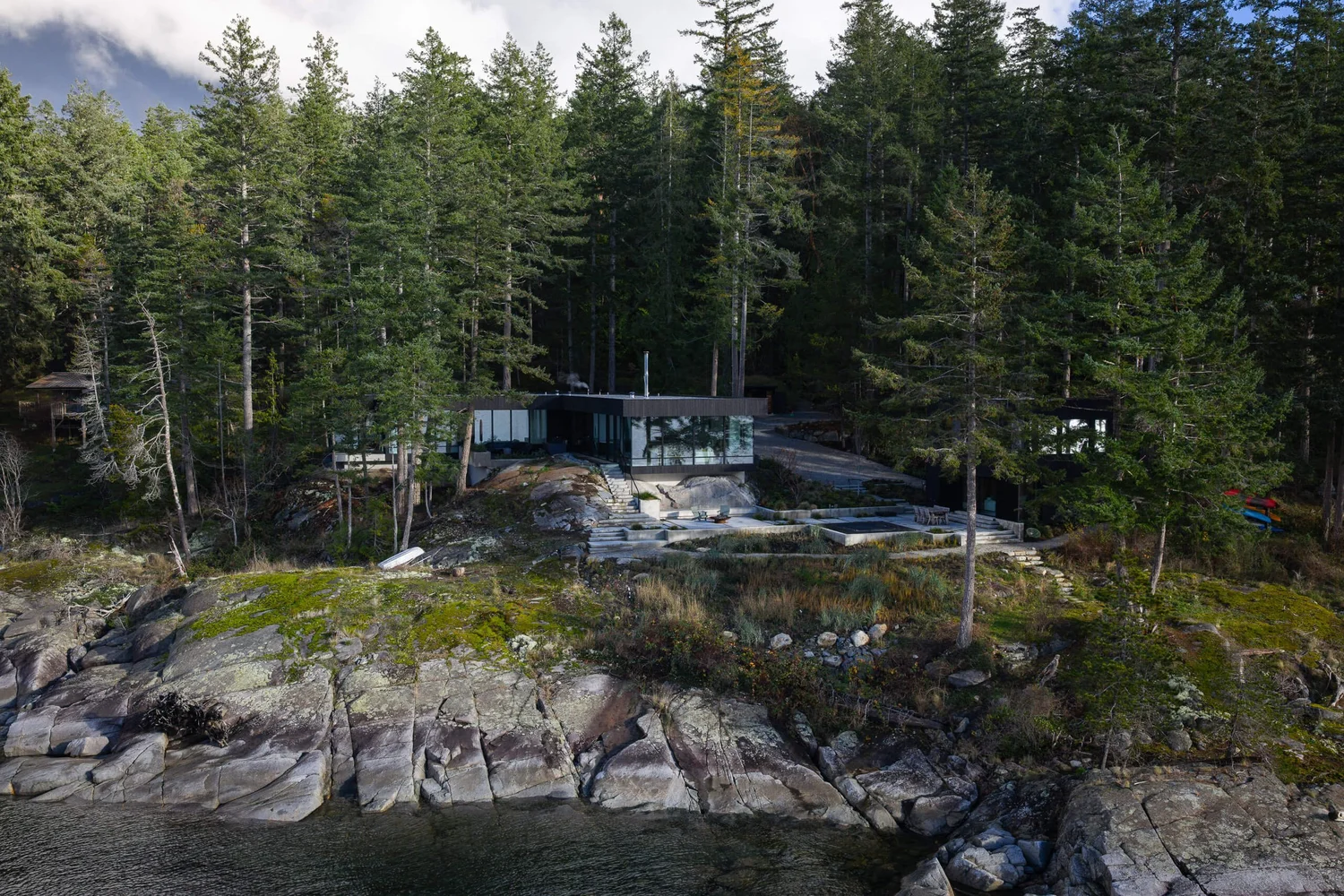 The house low on a forested rocky point, terraces stepping down toward the shore