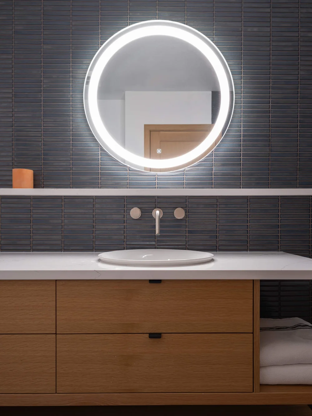 White basin under a round lit mirror, dark ribbed tile above pale timber drawers