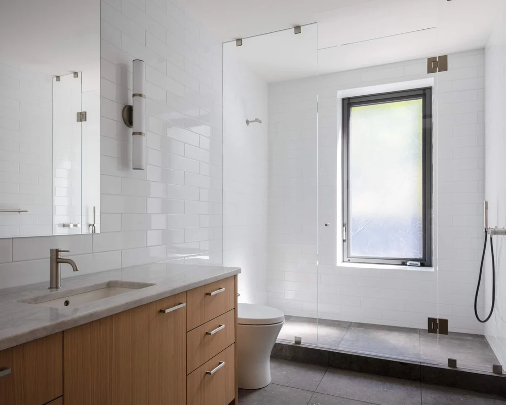 White tiled bathroom with a wood vanity, a glass shower and a window to the trees