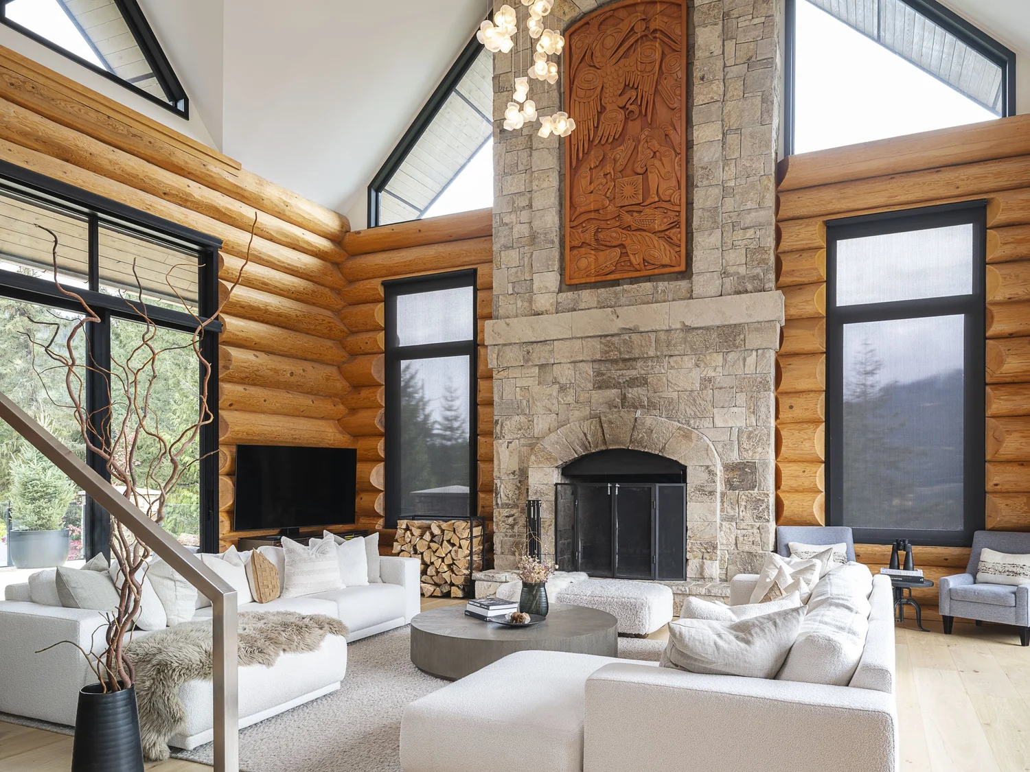 The great room under a vaulted log ceiling, a stone chimney and carved panel between tall windows