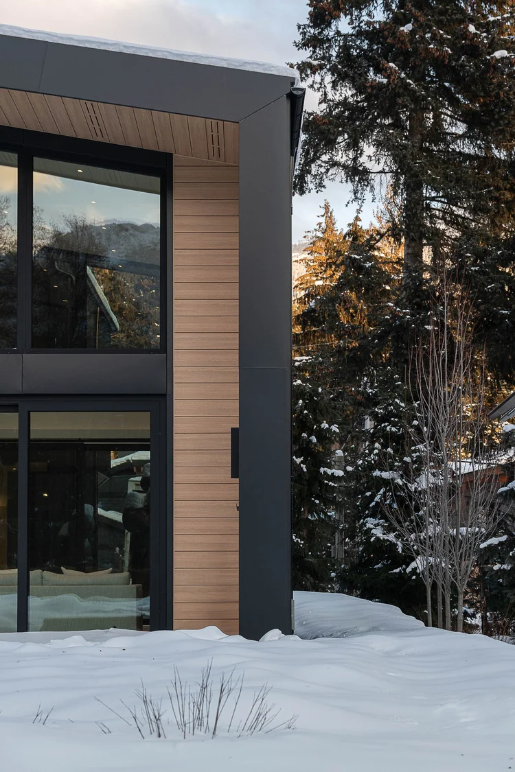 Cedar and dark metal exterior corner with glazing, snow and conifers in low sun