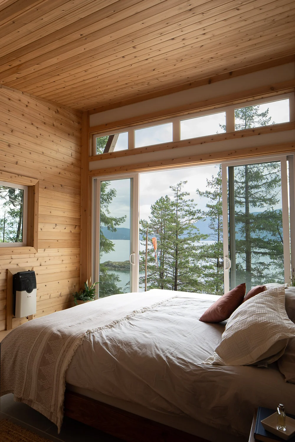 Bedroom lined in wood boards, windows and clerestory glazing onto trees and the water