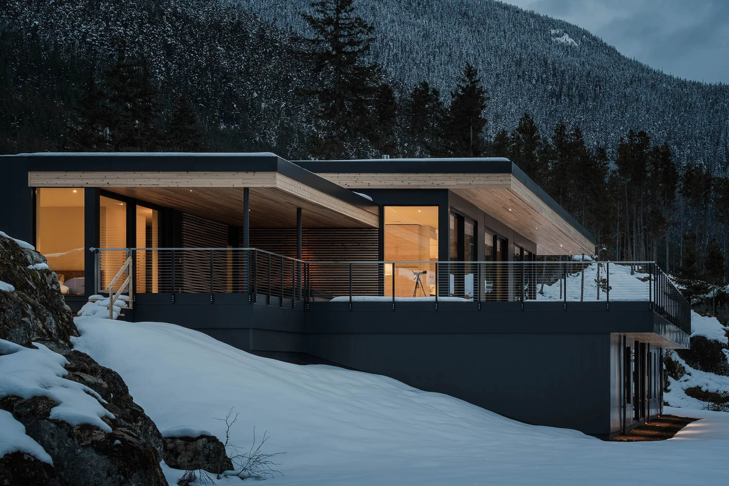 The house at dusk from below the rock, warm interior light along the dark elevation