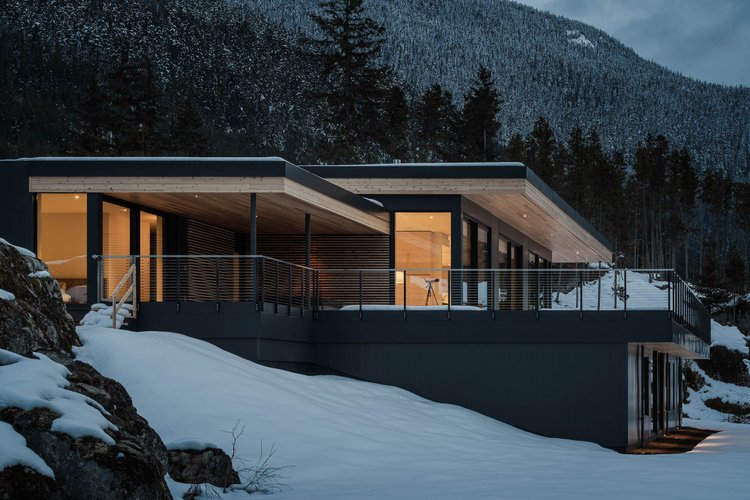 The house at dusk from below the rock, warm interior light along the dark elevation