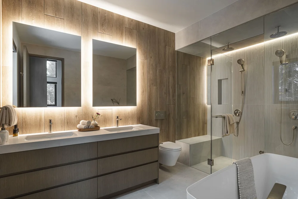 Two backlit mirrors over a long double vanity, glass shower and a tub at right